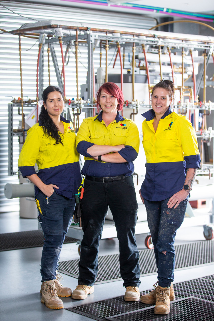 Female apprentice plumbers with a selection of pipes on a wall in the background.